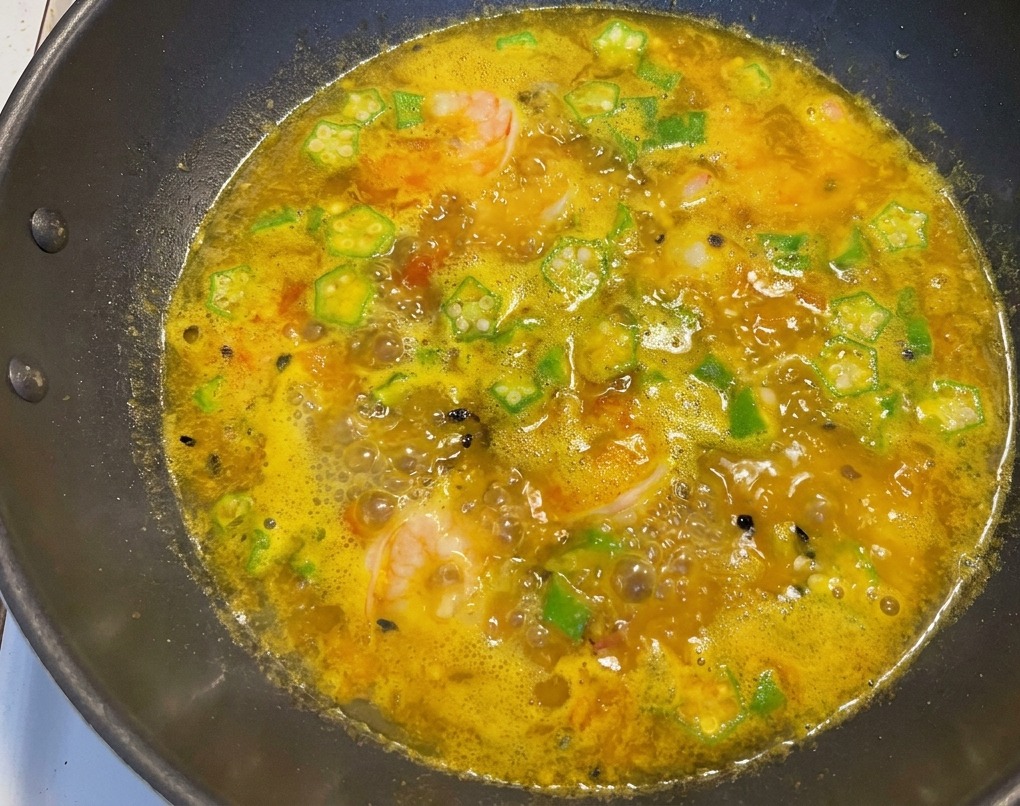 A dark cooking pan filled with bubbling golden soup, dotted with green okra slices, pink cooked shrimp, and dark passion fruit seeds.