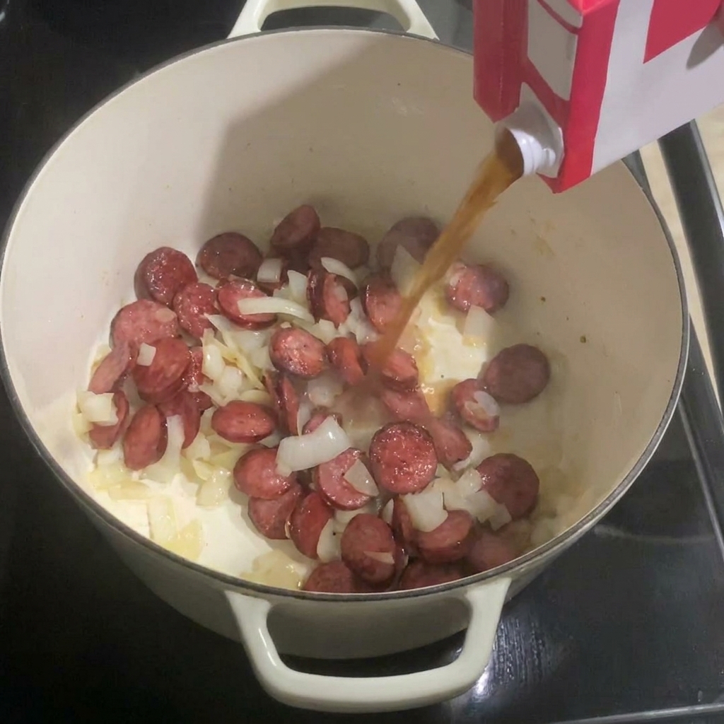 Pouring chicken broth from a carton into a white pot containing sliced sausage and cooked onions.