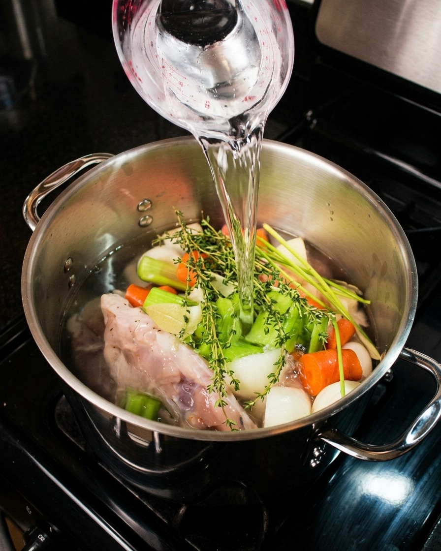 Clear water being poured from a measuring cup over raw chicken, carrots, celery, onions, and fresh thyme in a stainless steel stock pot.