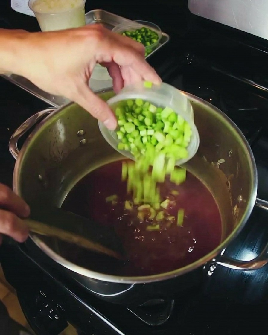 A hand pouring diced yellow onions from a small container into a large pot containing a bubbling dark brown roux.