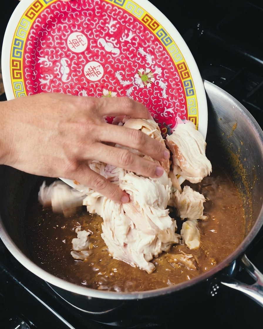 A hand sliding a mound of shredded, cooked white chicken meat from a red patterned plate into a large pot of dark, rich gumbo.