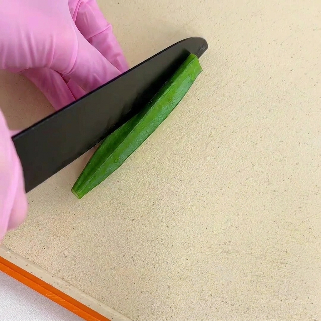 Gloved hands slicing a blanched green okra pod perfectly in half lengthwise with a black knife.