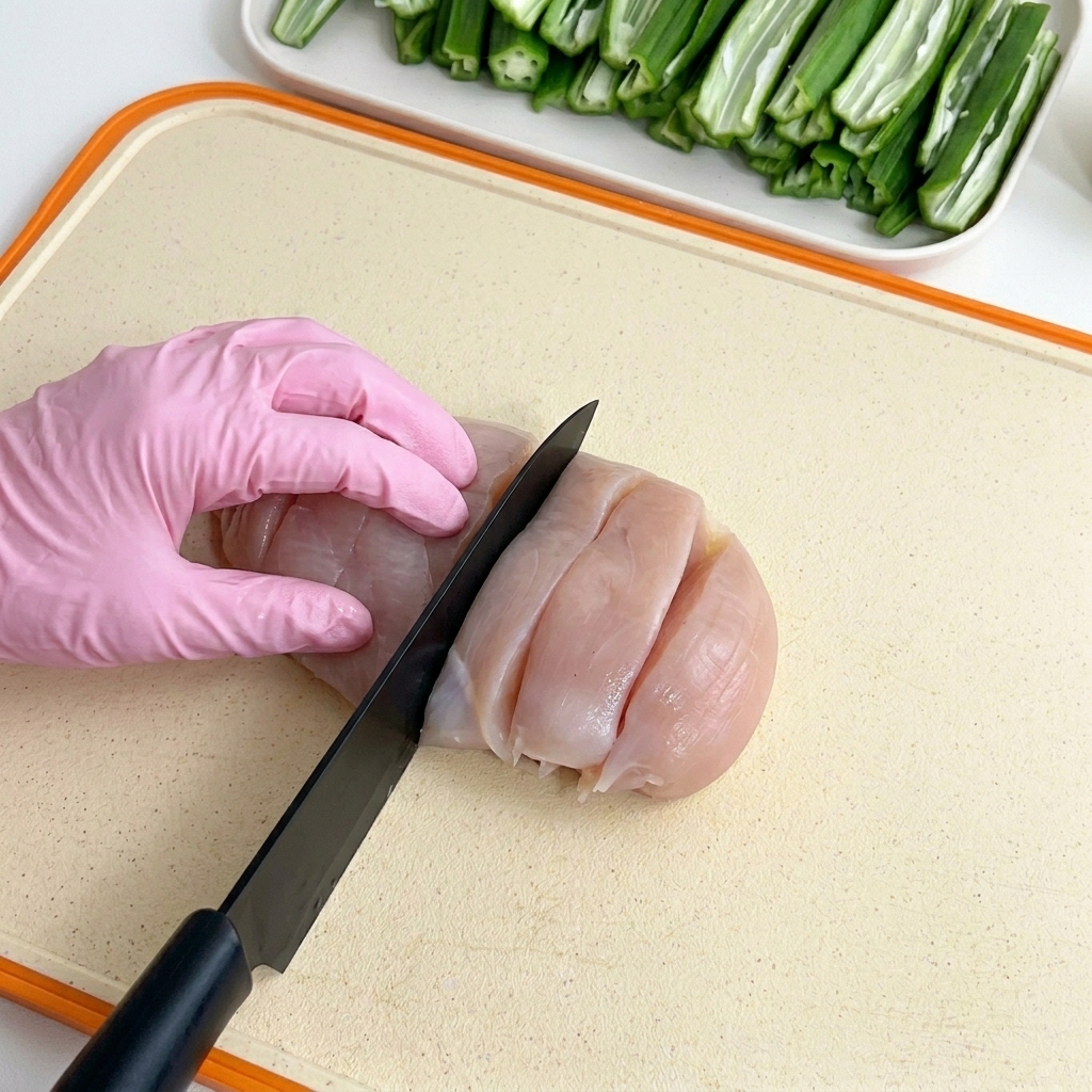 Gloved hands slicing a raw chicken breast into thick sections with a black knife on a cutting board, with prepared okra in the background.