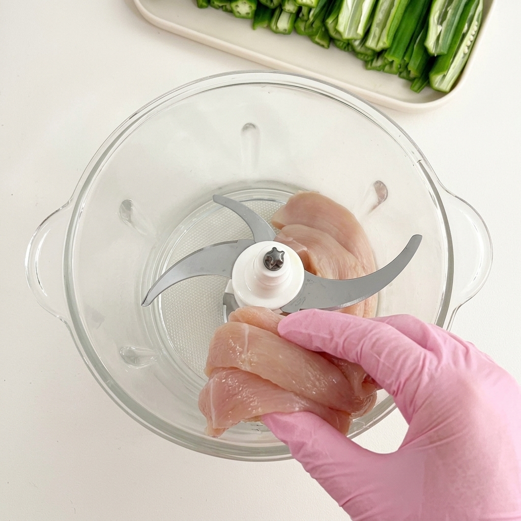 Gloved hand dropping slices of raw chicken breast into a glass food processor bowl fitted with a sharp metal blade.