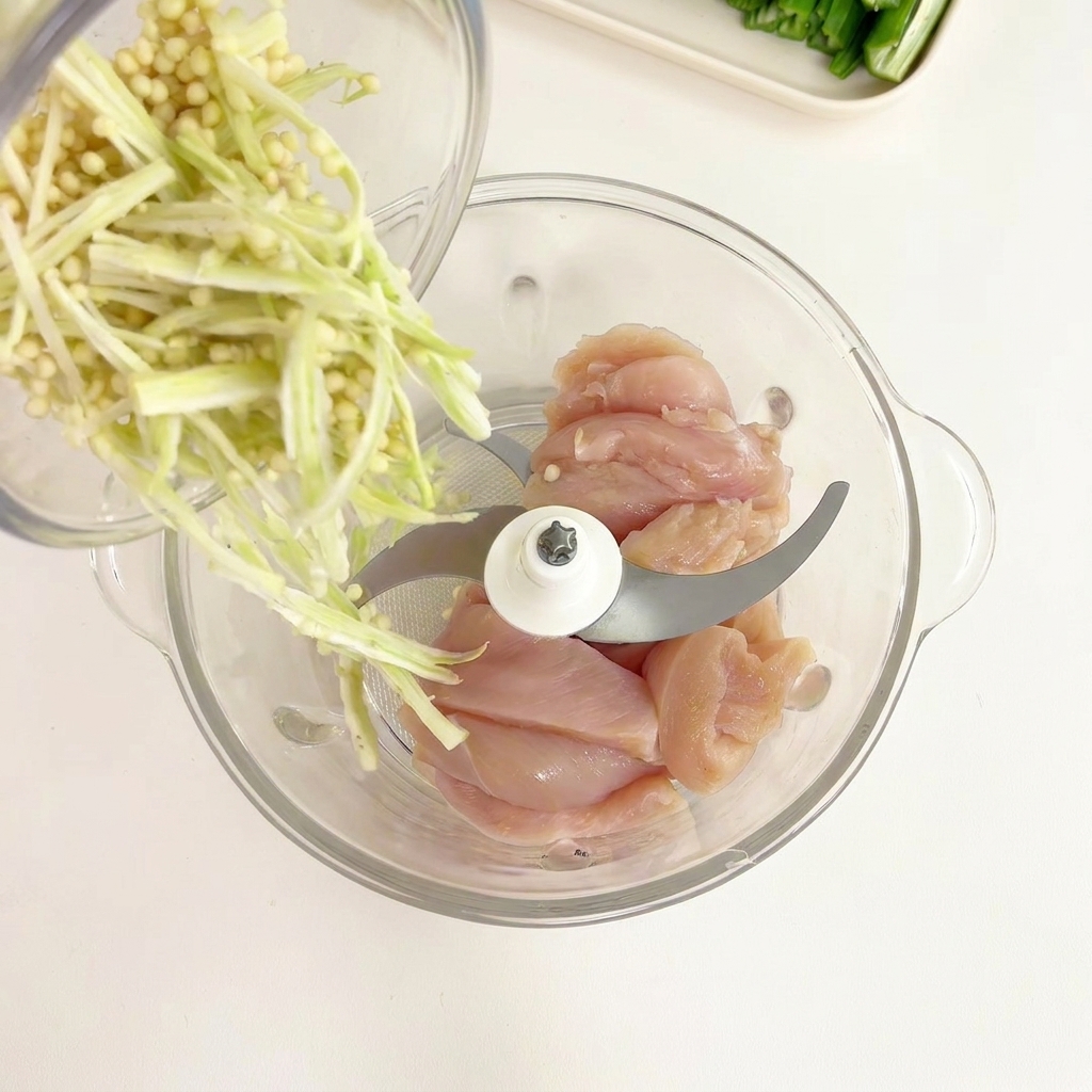 Okra pith and seeds being poured from a glass bowl into a food processor containing chunks of raw chicken breast.
