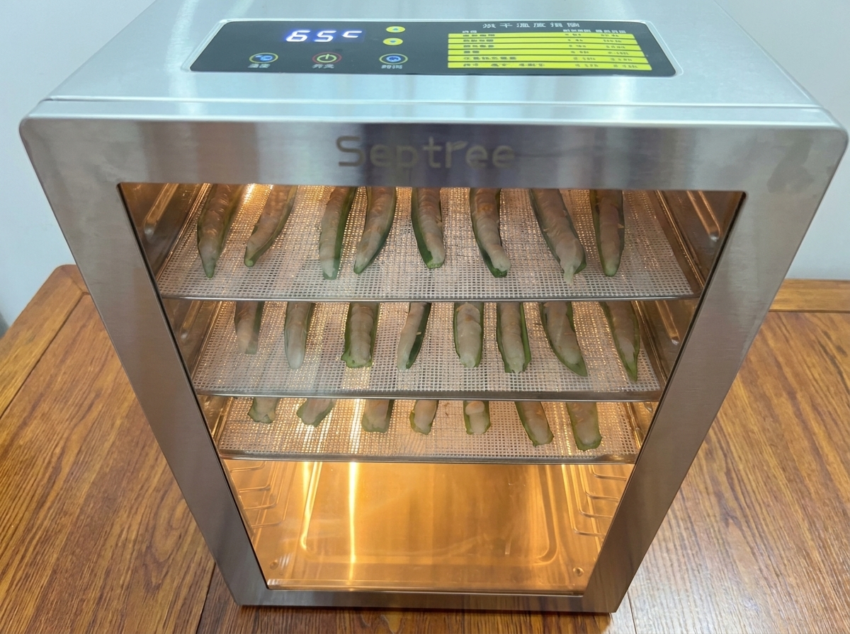 Mesh trays loaded with stuffed okra being placed into a brightly lit stainless steel food dehydrator.