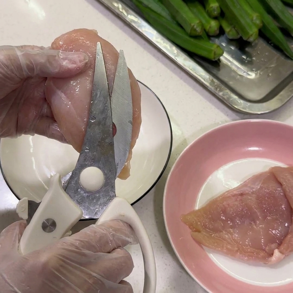 Gloved hands using kitchen shears to snip white fat off of a raw chicken breast over a plate.