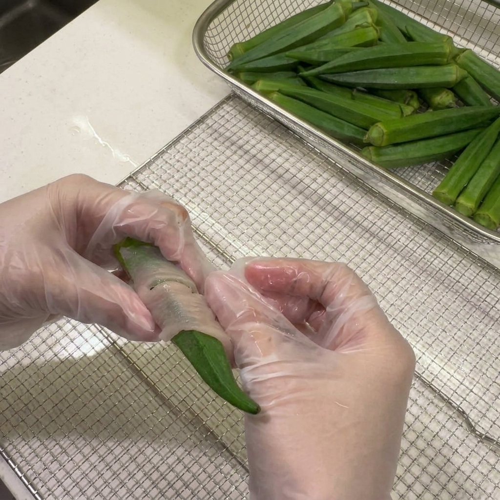 Gloved hands firmly wrapping a thin strip of raw chicken breast around a fresh green okra pod over a wire mesh dehydrator tray.