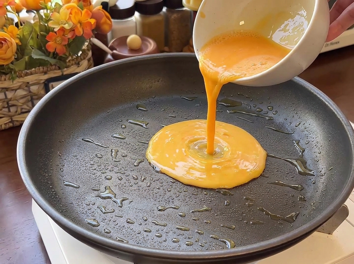Smooth yellow egg liquid being poured from a bowl into a large, dark non-stick frying pan with visible oil droplets.