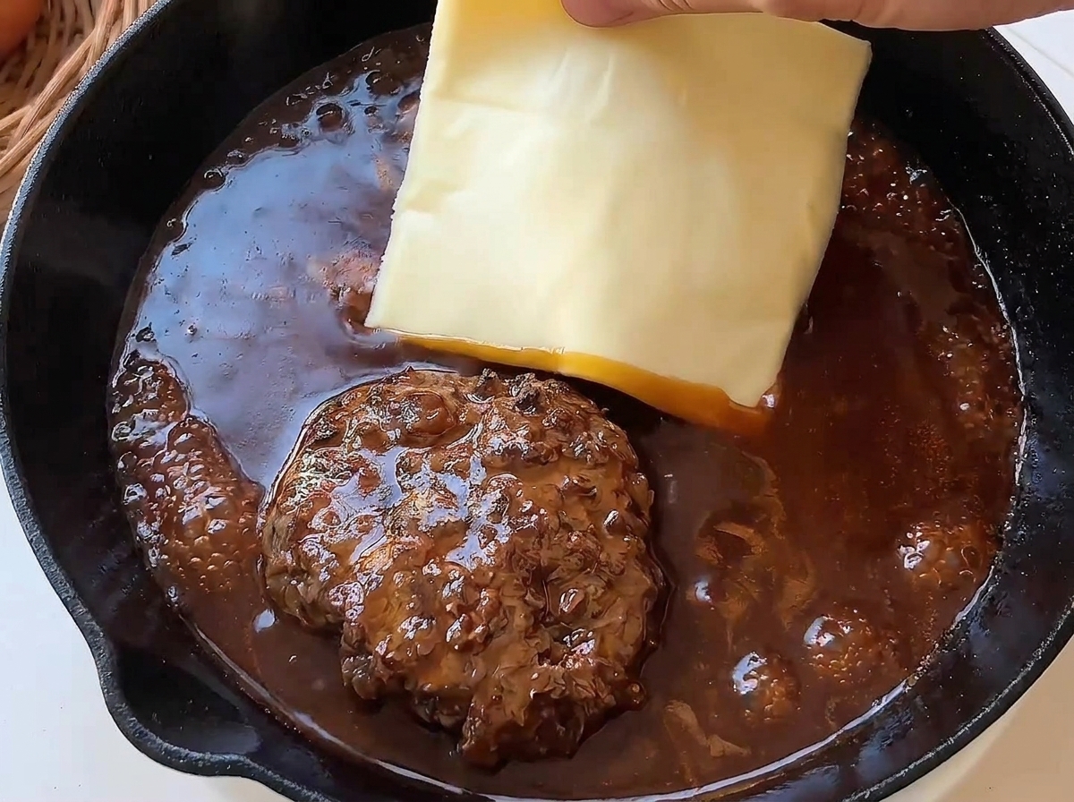 A hand placing a square slice of yellow cheese onto a hot beef patty simmering in a rich brown sauce.