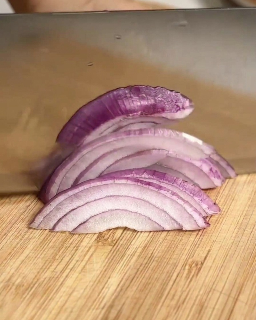 A large knife slicing a red onion into strips on a bamboo cutting board.