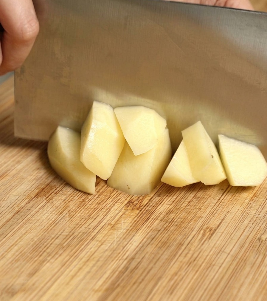 A metal cleaver chopping a peeled yellow potato into chunks on a wooden board.
