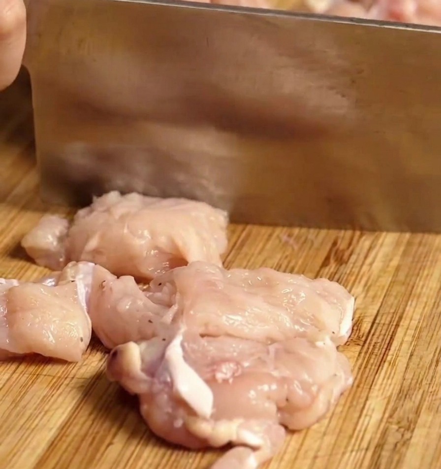 A knife slicing raw chicken breast into small chunks on a wooden cutting board.