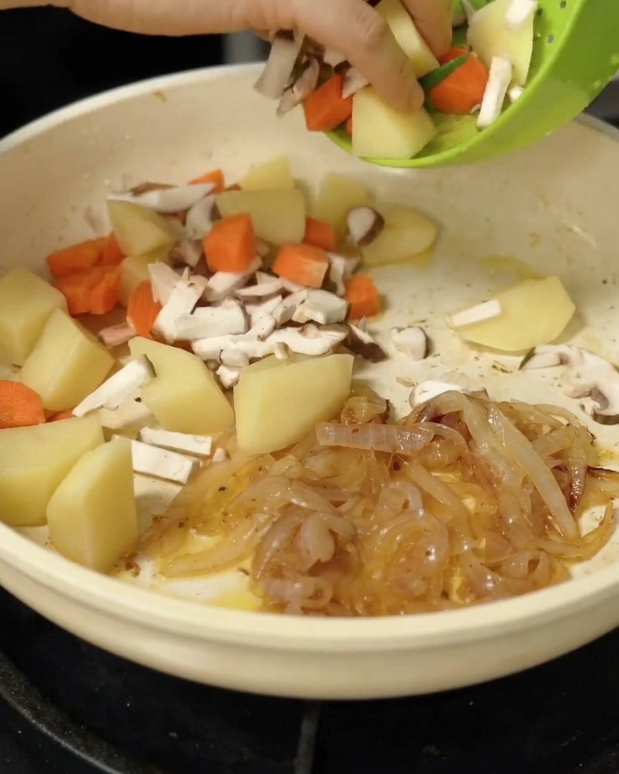 Adding chunks of potatoes, carrots, and chopped shiitake mushrooms to the sautéed onions in a skillet.