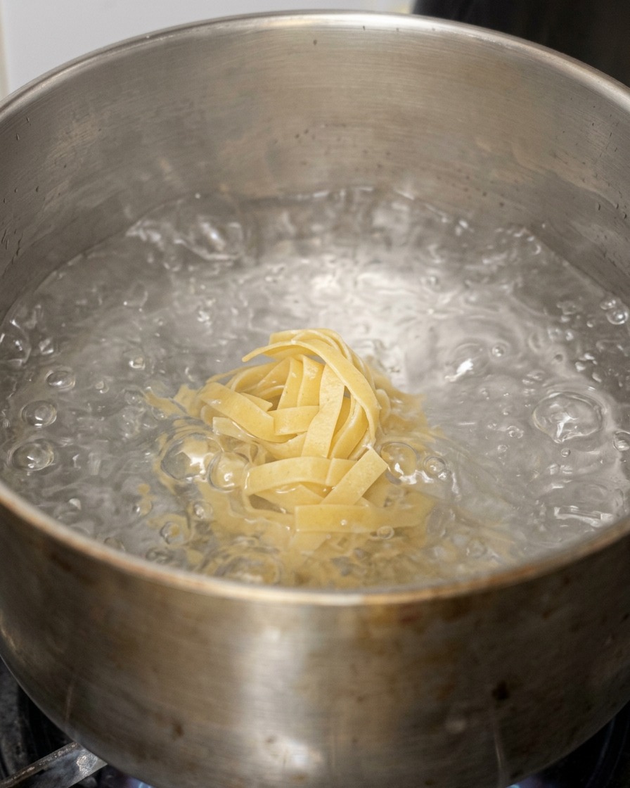 A nest of flat, wide pasta noodles being dropped into a metal pot of rolling boiling water.