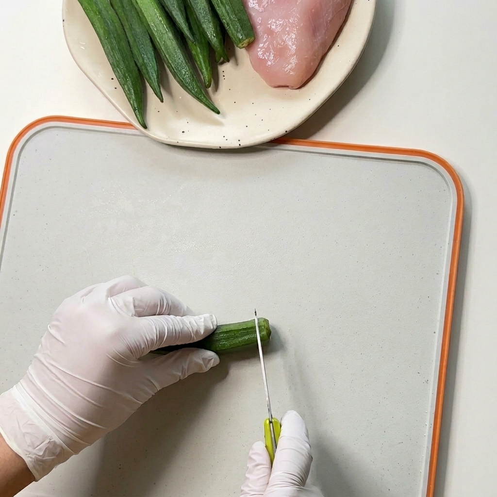 Gloved hands using a small knife to cut the ends off fresh green okra pods on a white cutting board.