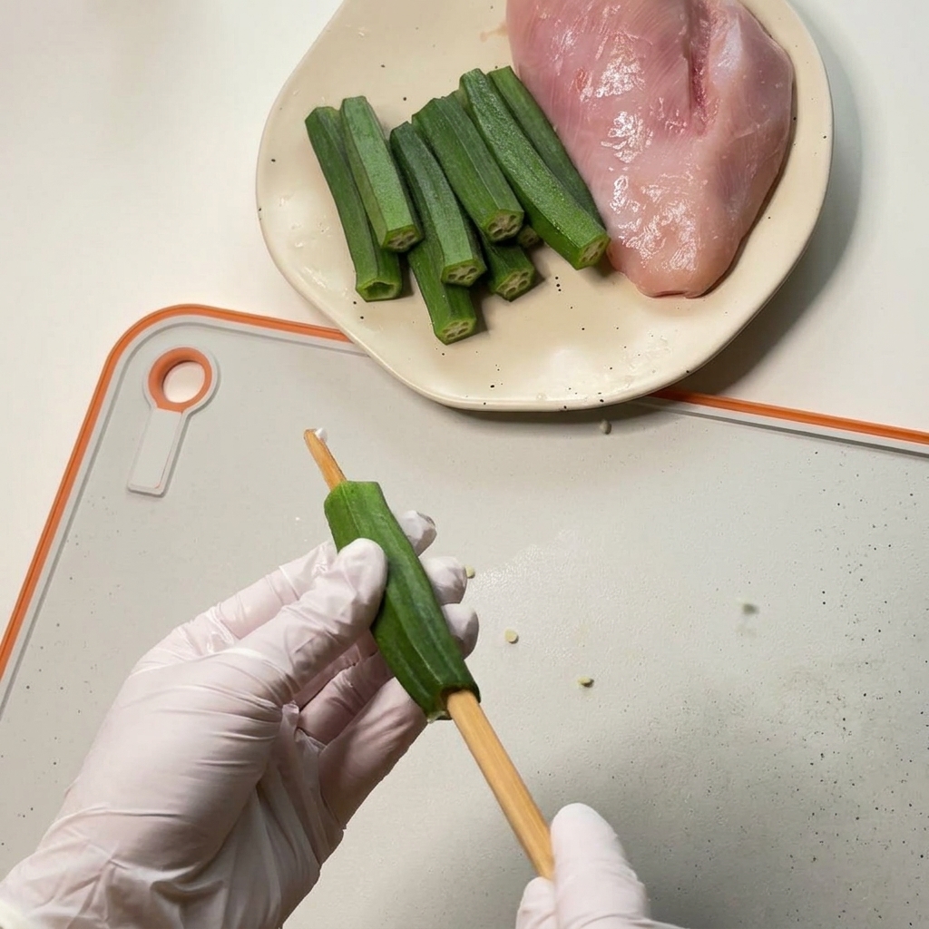 Gloved hands hollowing out the center of a green okra pod using a thick wooden chopstick over a white cutting board.