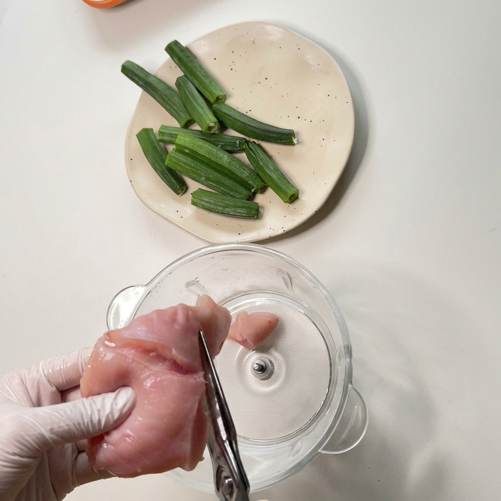 Gloved hands using kitchen scissors to cut a piece of raw chicken breast over a glass food processor bowl.
