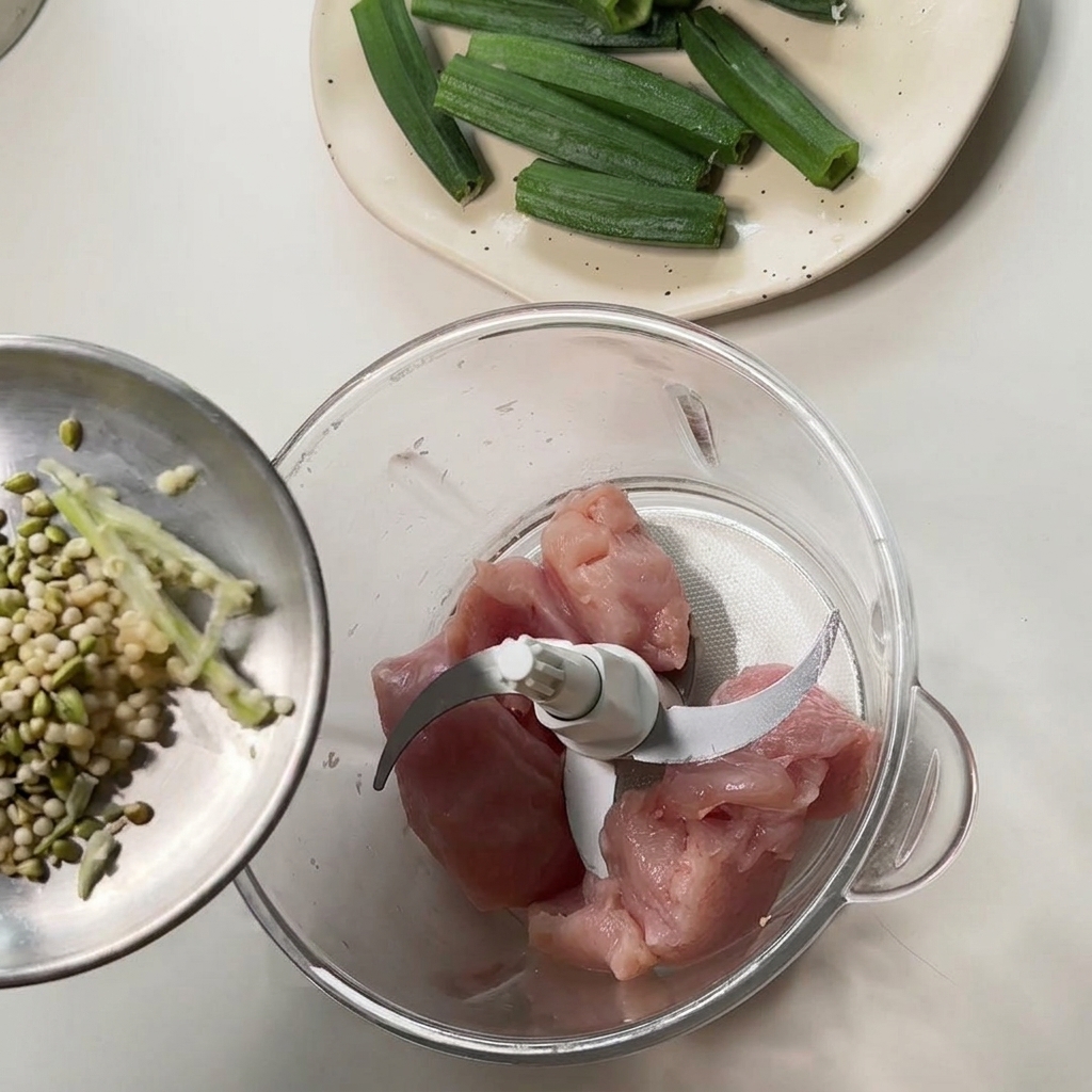 Pouring okra seeds and scraps from a small metal dish into a food processor containing raw chicken chunks.