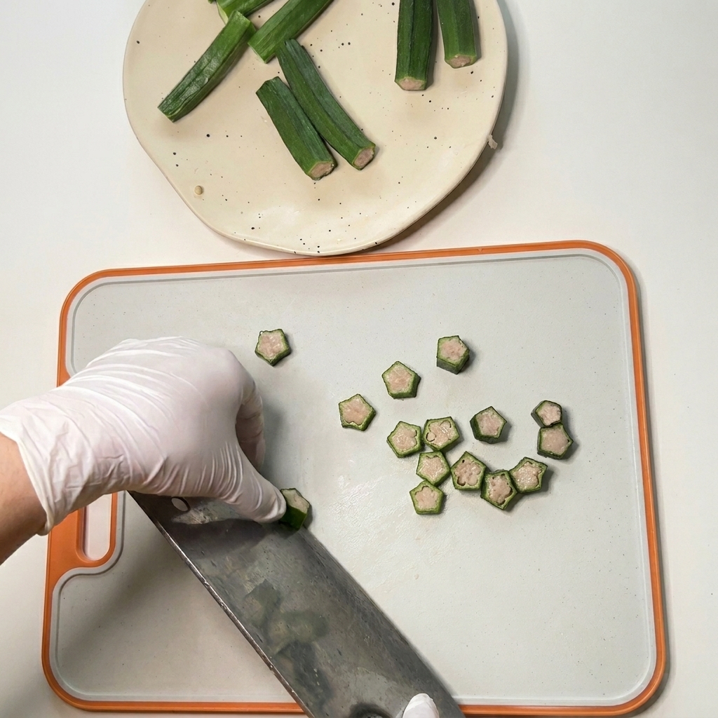 A gloved hand using a cleaver to slice raw, chicken-stuffed okra into small star shapes on a cutting board.