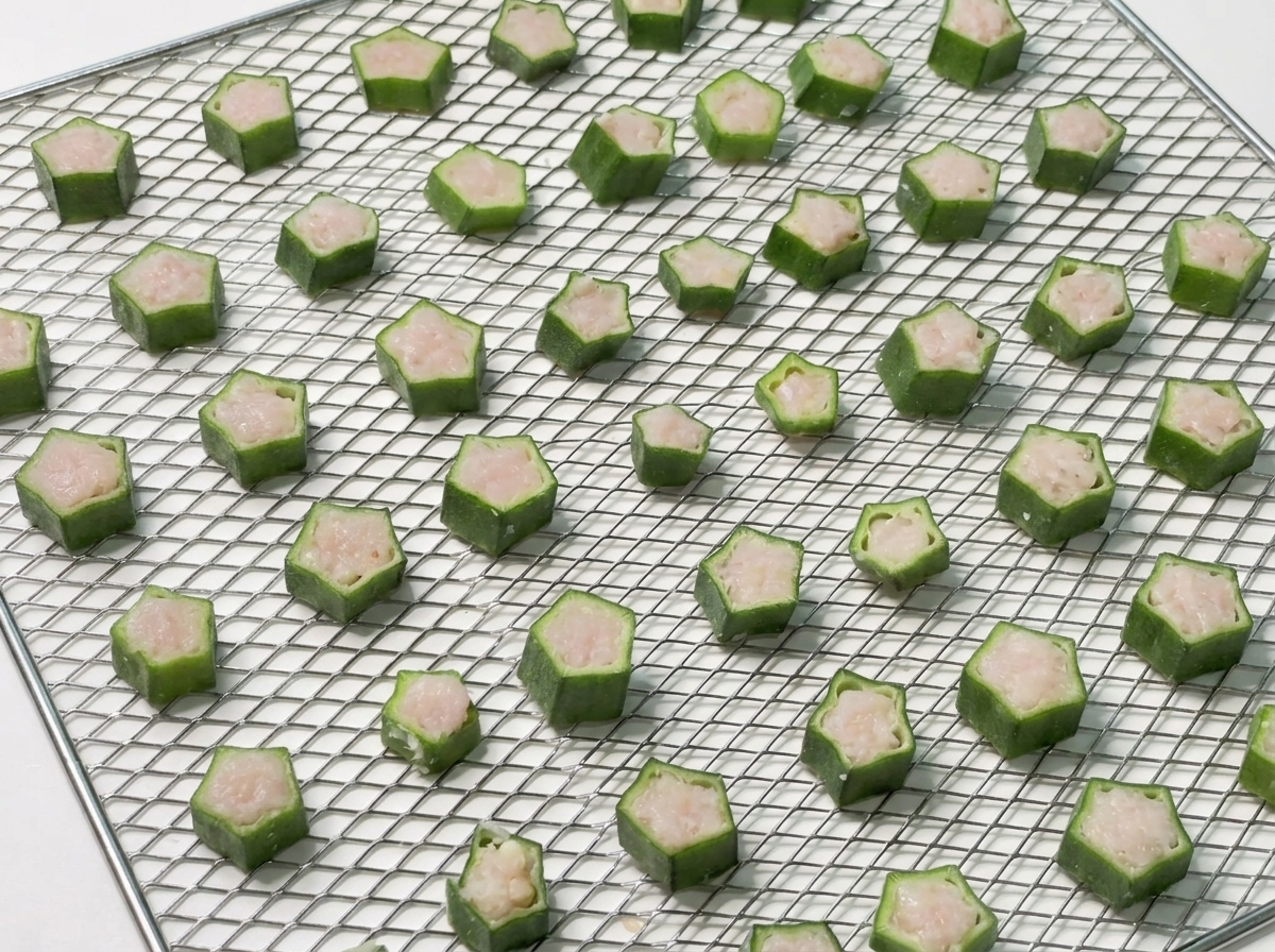 A metal mesh tray perfectly organized with raw, chicken-stuffed okra slices ready for the food dehydrator.