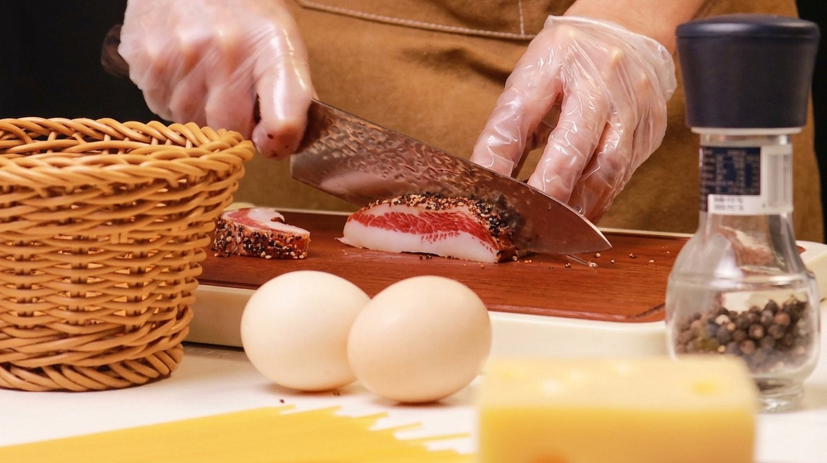 Hands wearing clear plastic gloves slicing a piece of pepper-crusted guanciale on a wooden cutting board.