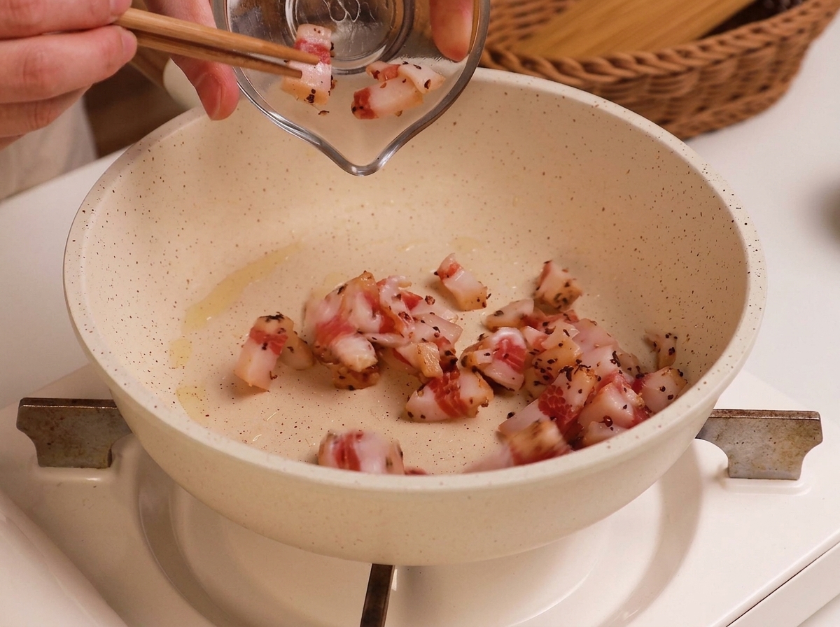 Diced raw guanciale being transferred from a small glass bowl into a lightly oiled white frying pan.