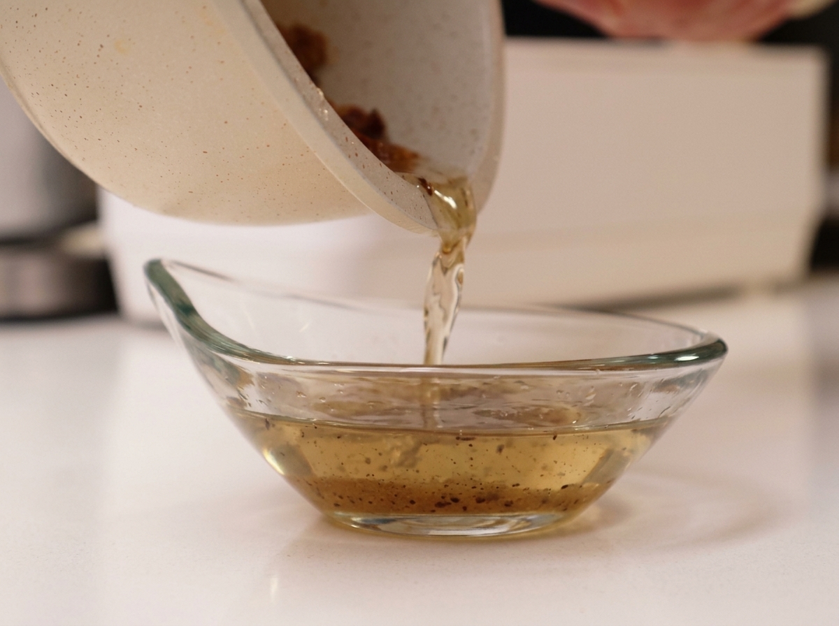 Pouring hot rendered pork fat from a white pan into a clear glass bowl.