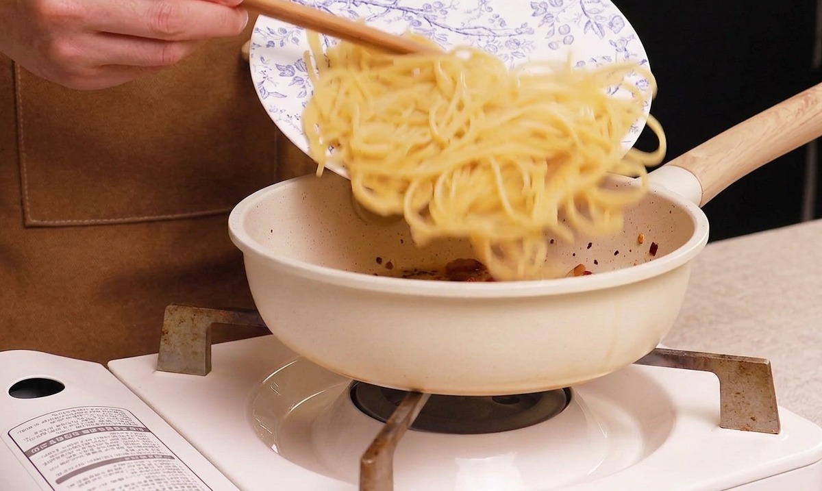 Sliding cooked spaghetti from a blue and white floral plate into a white frying pan containing crispy cooked guanciale.