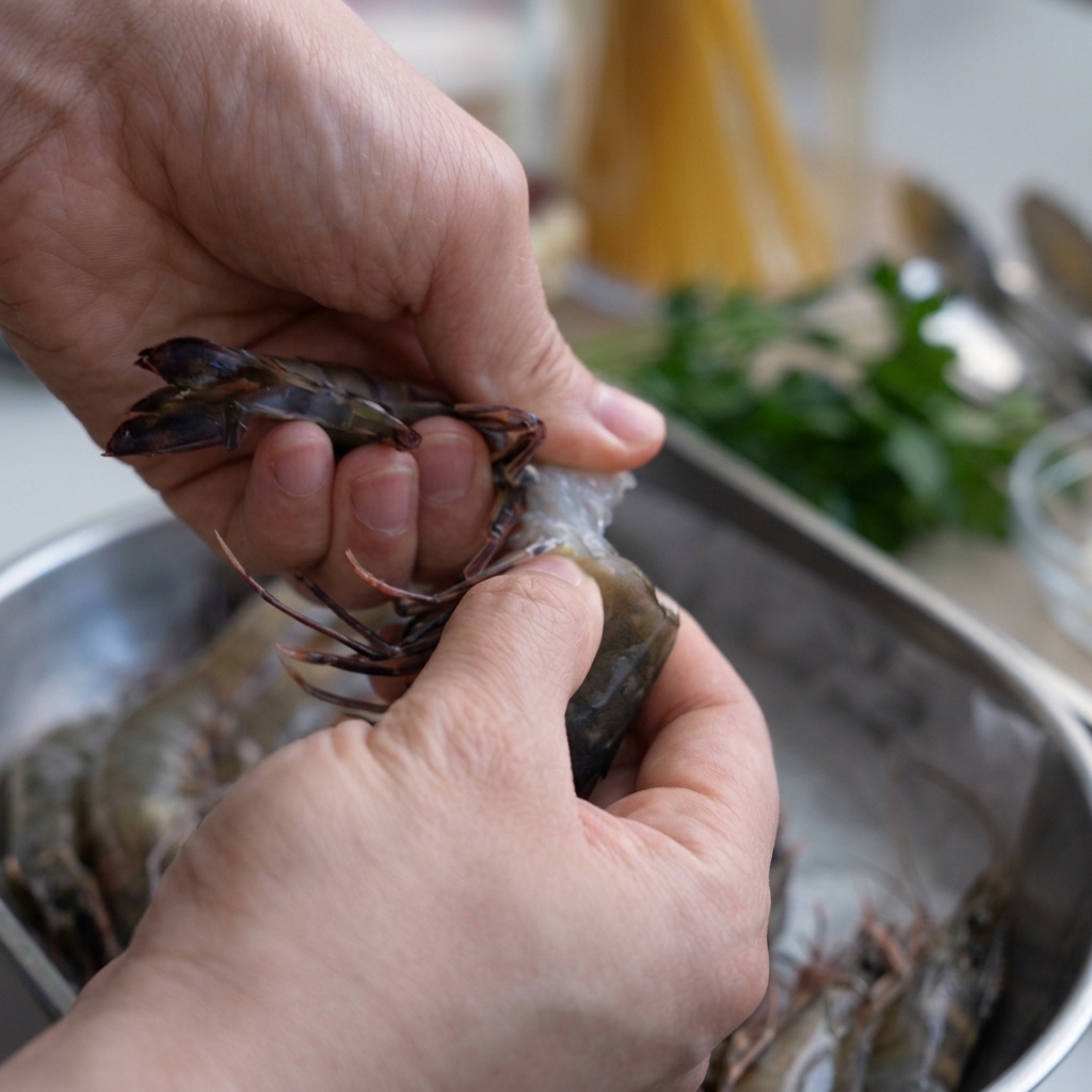 Hands peeling a raw tiger prawn over a stainless steel bowl, keeping the head intact for later use.