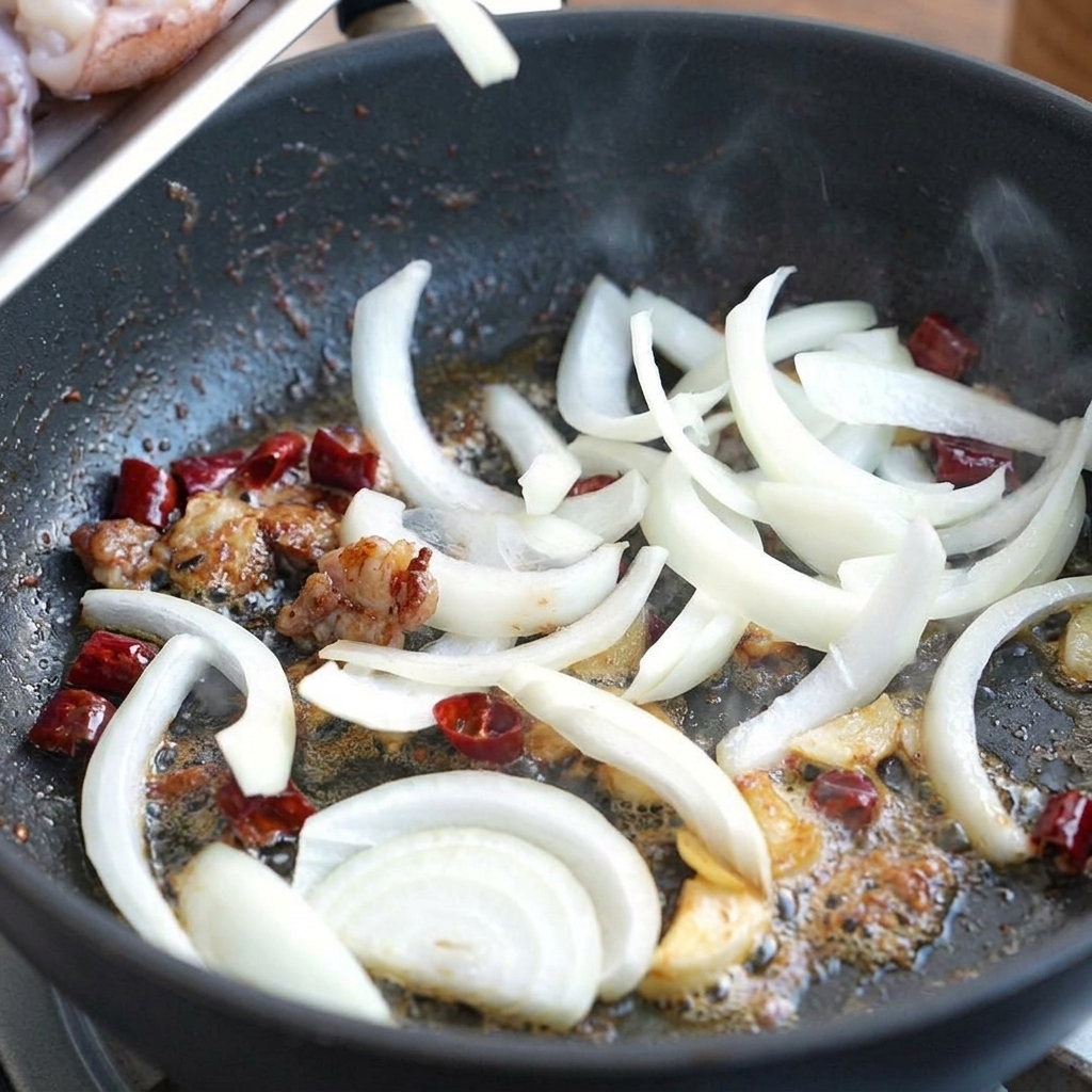 Sliced white onions being tossed in a pan with seasoned oil, dried chilies, and garlic.