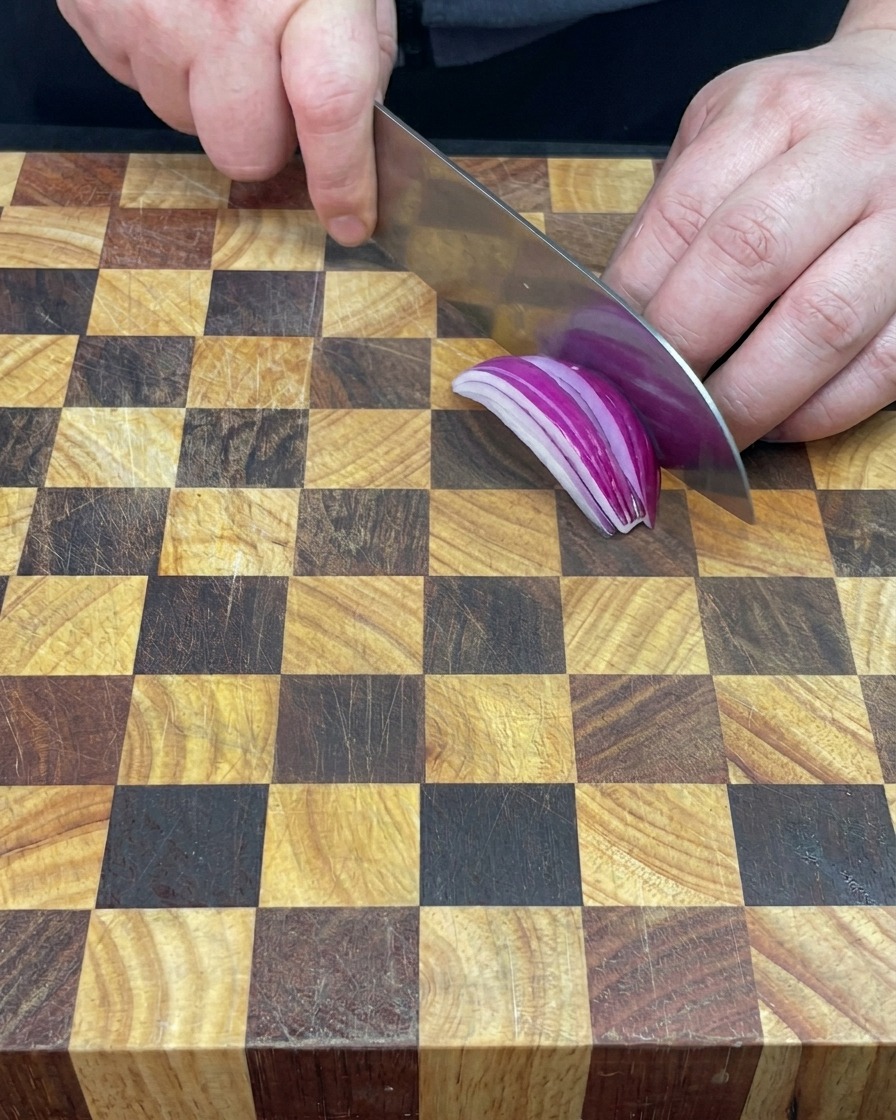 A hand holding a knife and dicing a bright purple red onion half on a wooden cutting board.