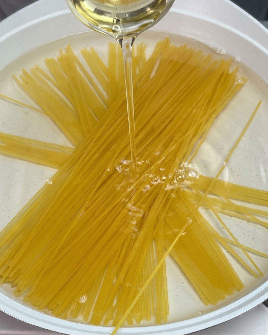 Dry spaghetti noodles submerged in a pot of water while a stream of cooking oil is poured over them.