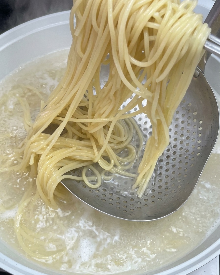 Cooked spaghetti noodles being lifted from a pot of boiling water using a metal skimmer.