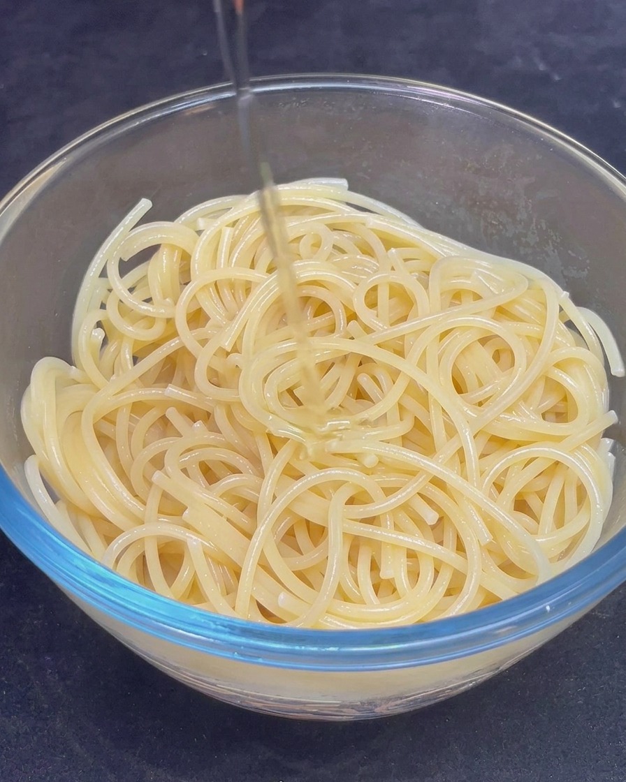 A stream of golden olive oil being poured over a glass bowl filled with cooked spaghetti.
