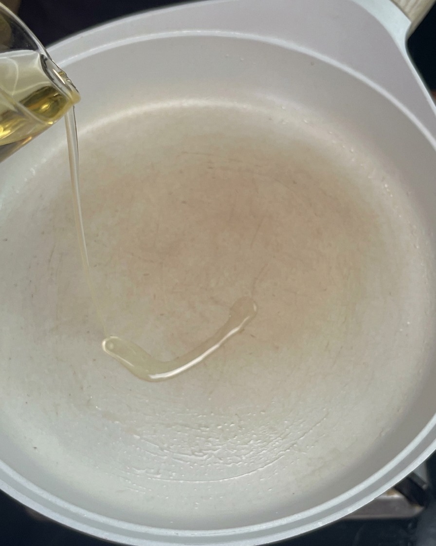 Clear cooking oil being poured from a small glass pitcher into a preheated empty white pan.