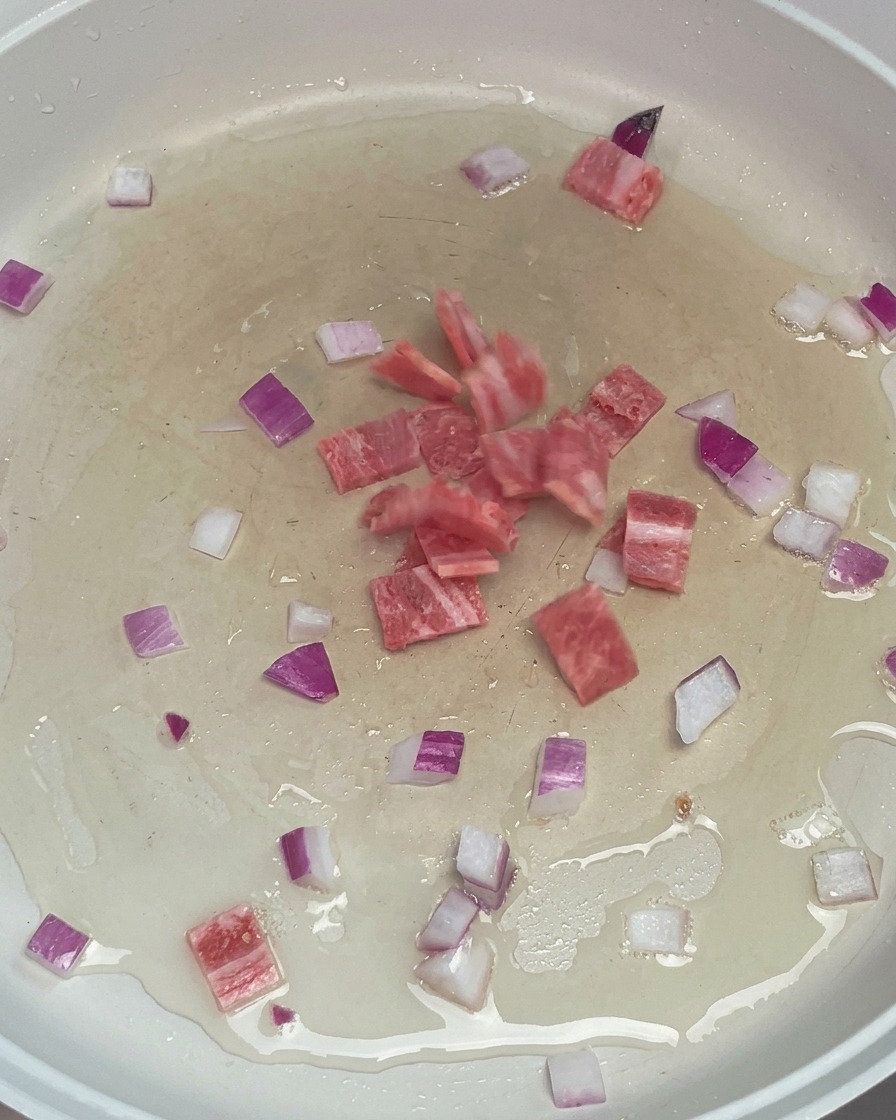 Diced bacon and chopped red onions sizzling in hot oil in a white frying pan.
