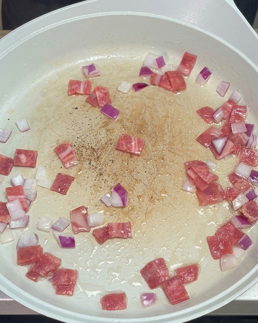Ground black pepper sprinkled over sautéing diced bacon and red onions in a white pan.