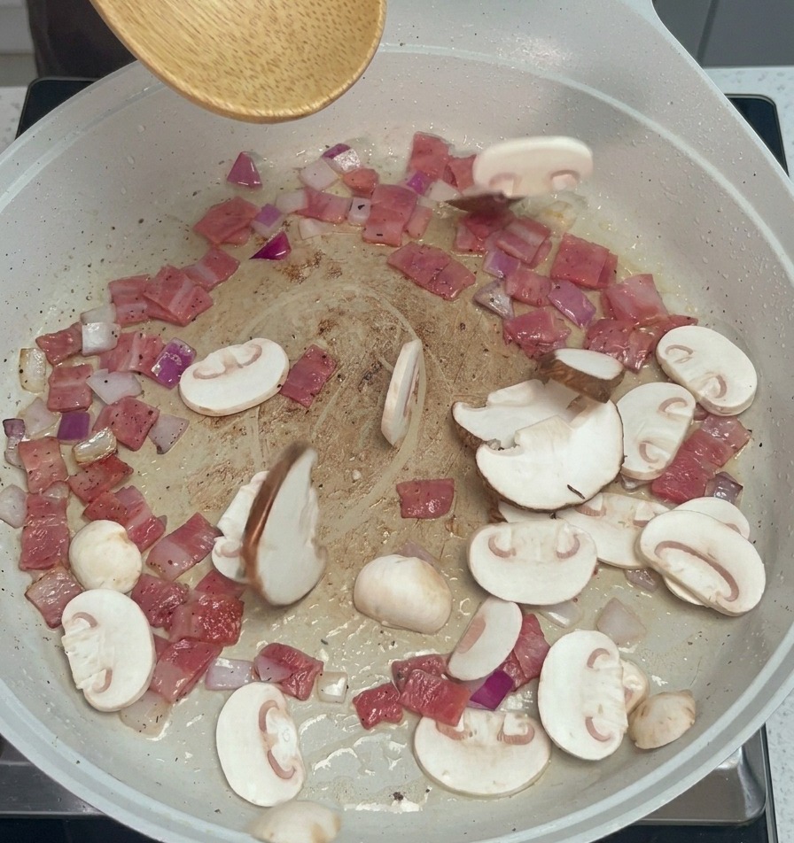 Sliced button and shiitake mushrooms being tossed into a pan with sautéed bacon and onions using a wooden spoon.