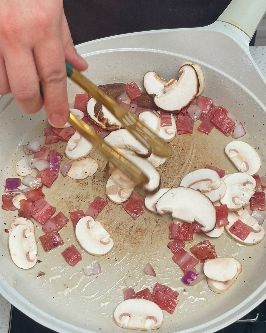 A hand using tongs to stir sliced mushrooms, diced red onions, and diced bacon in a white pan.