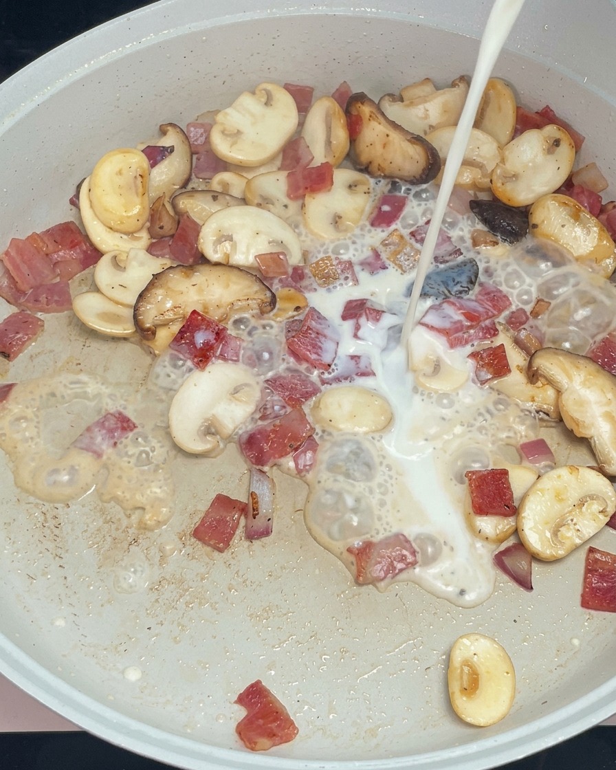 White milk being poured into a pan with sautéed mushrooms, bacon, and red onions.
