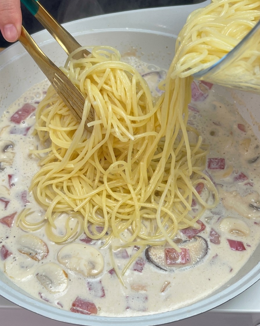 Tongs placing cooked spaghetti into a simmering creamy sauce with mushrooms and bacon.