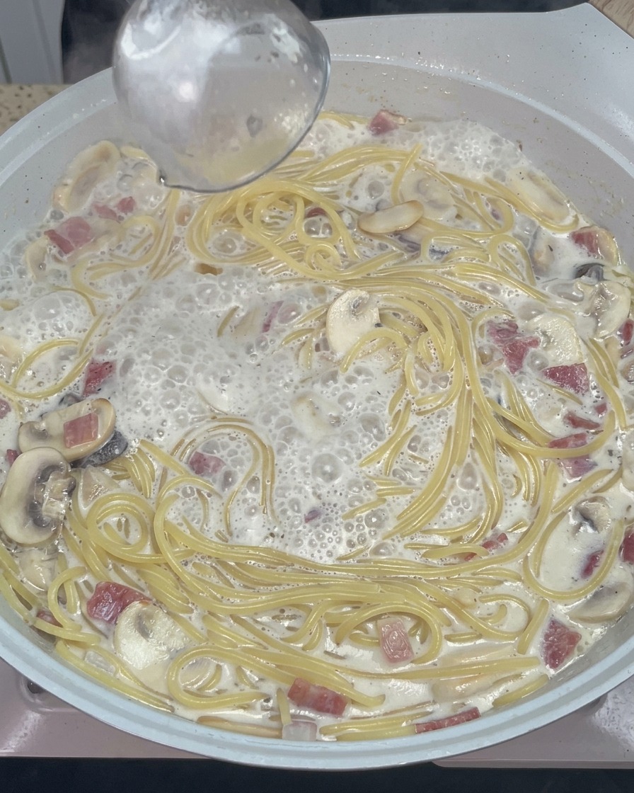A wooden spoon stirring creamy spaghetti with melted cheese and seasoning in a warm pan.