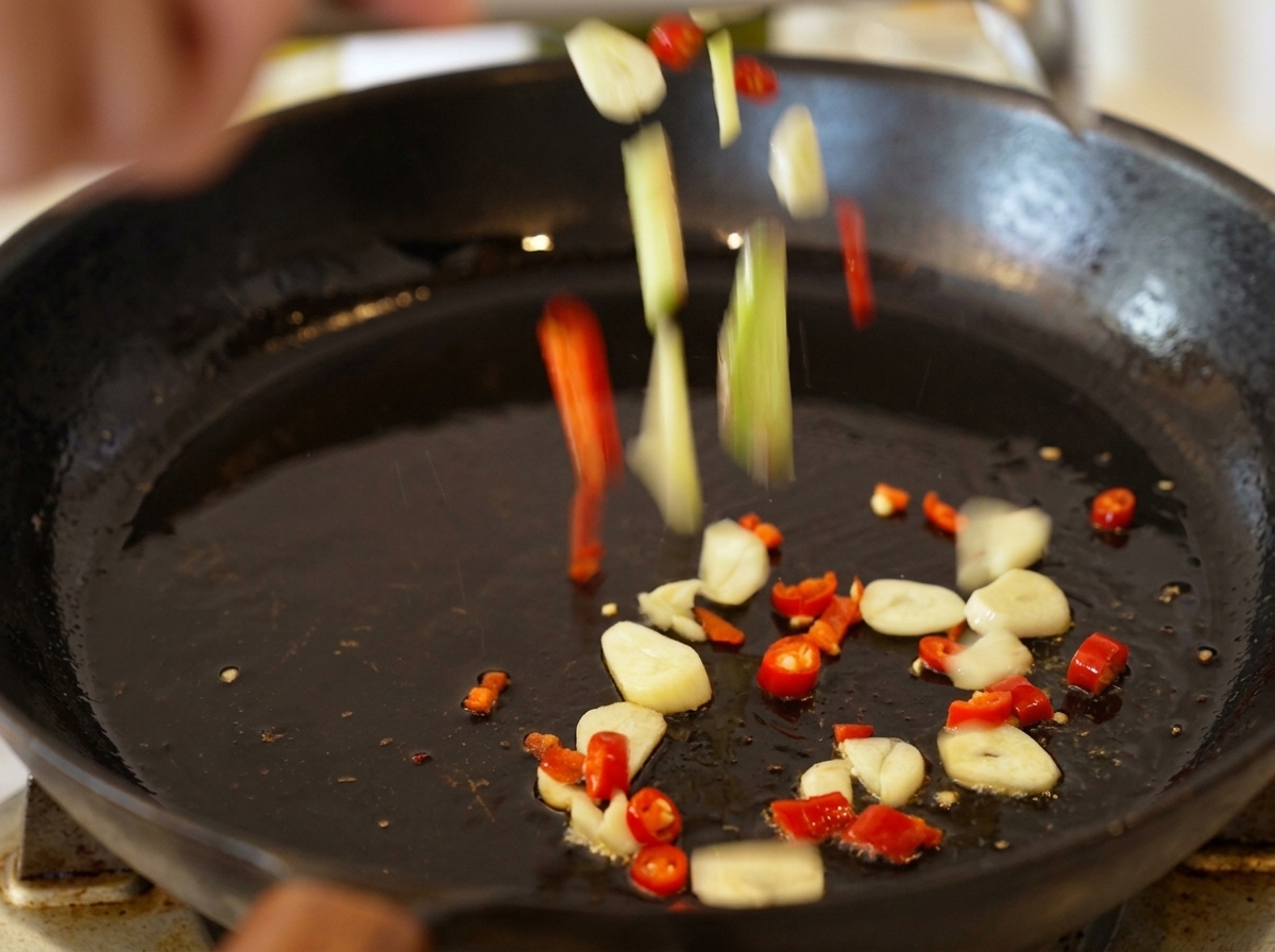 Sliced garlic and pieces of red chili falling into a black skillet with shimmering hot olive oil.