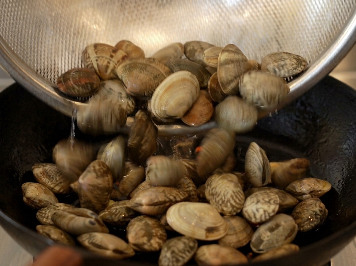 A batch of cleaned raw clams being poured from a metal mesh strainer into a hot cast iron skillet.