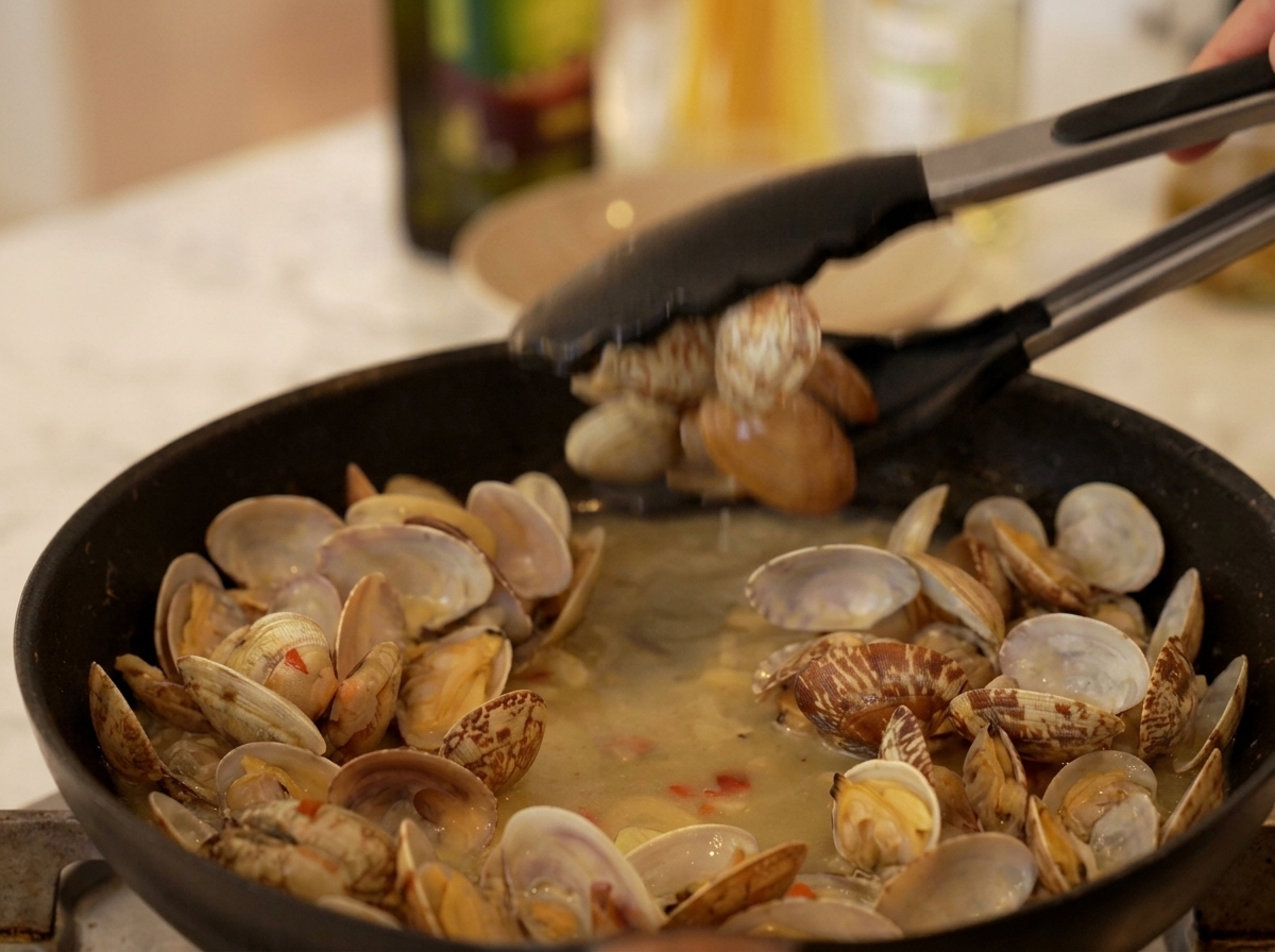 Black kitchen tongs picking up open, cooked clams from a simmering broth in a black skillet.
