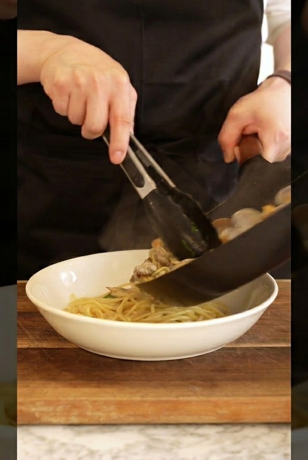 A cook wearing a black apron using tongs to transfer hot clam spaghetti from a pan into a white bowl.