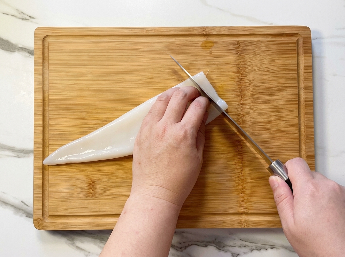 A person slicing a raw white squid tube into rings on a bamboo cutting board.