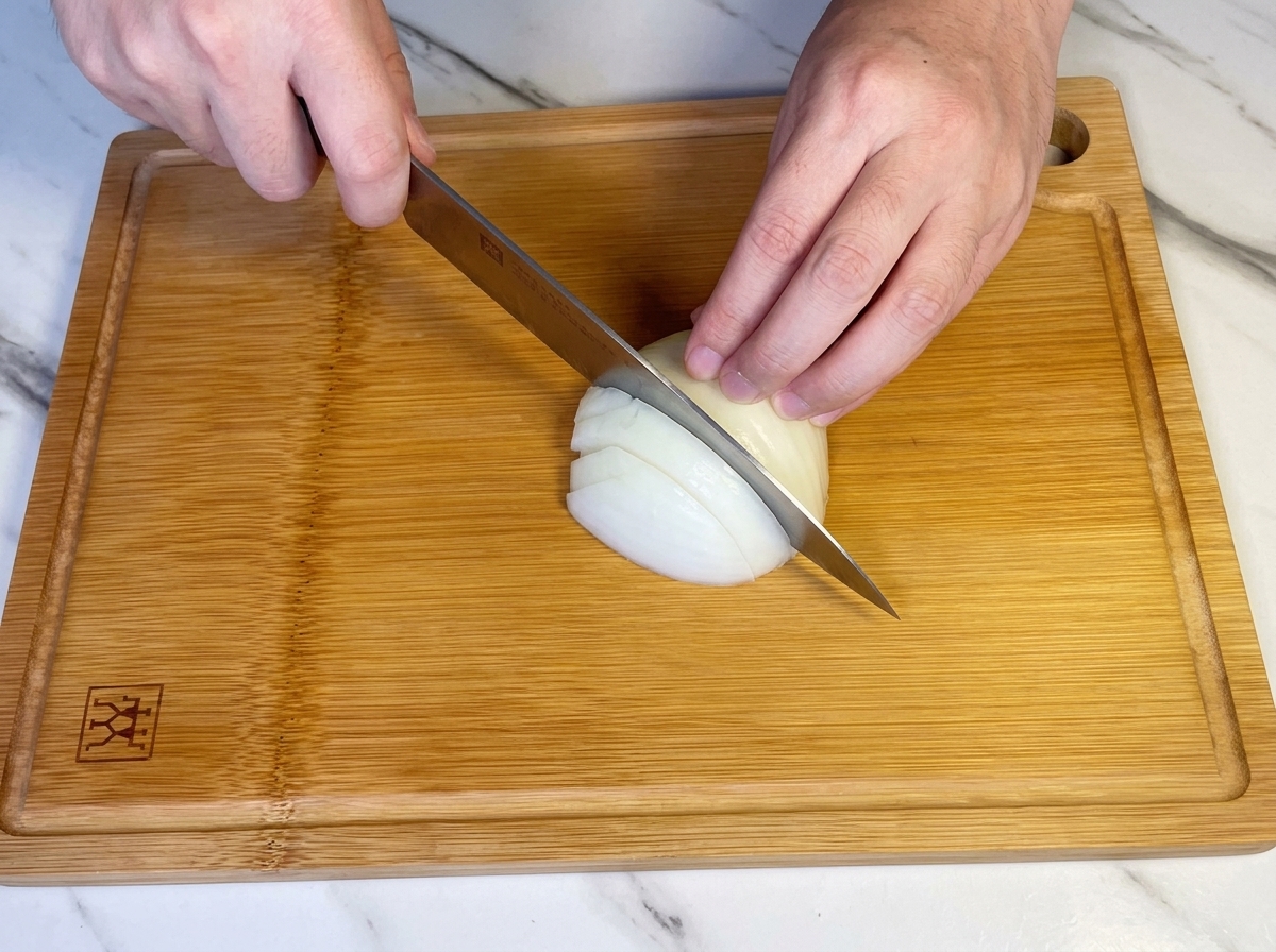 Hands using a chefs knife to dice half of a white onion on a wooden cutting board.