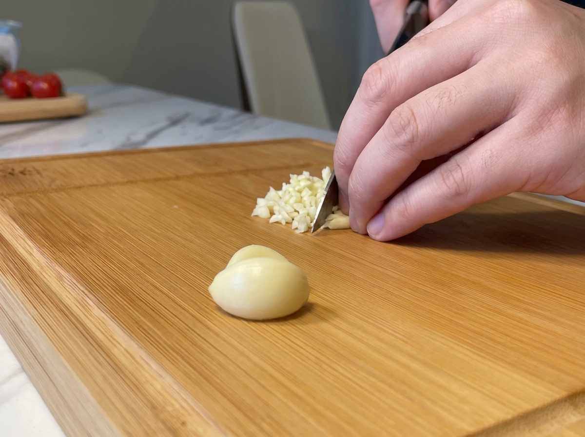 Finely mincing fresh garlic cloves into small pieces with a knife on a wooden chopping block.